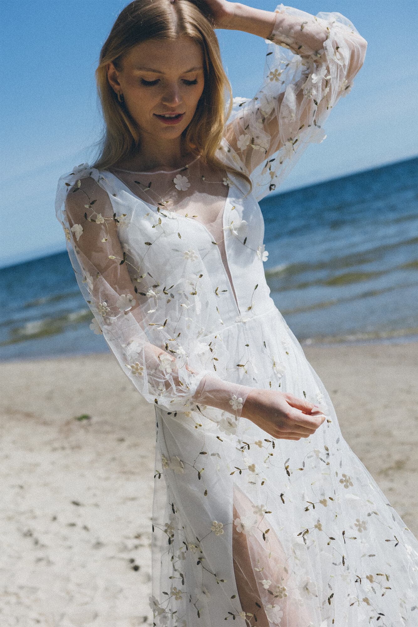 Suknia ślubna z przezroczystymi rękawami ozdobionymi kwiatowymi haftami, widoczna na plaży.
