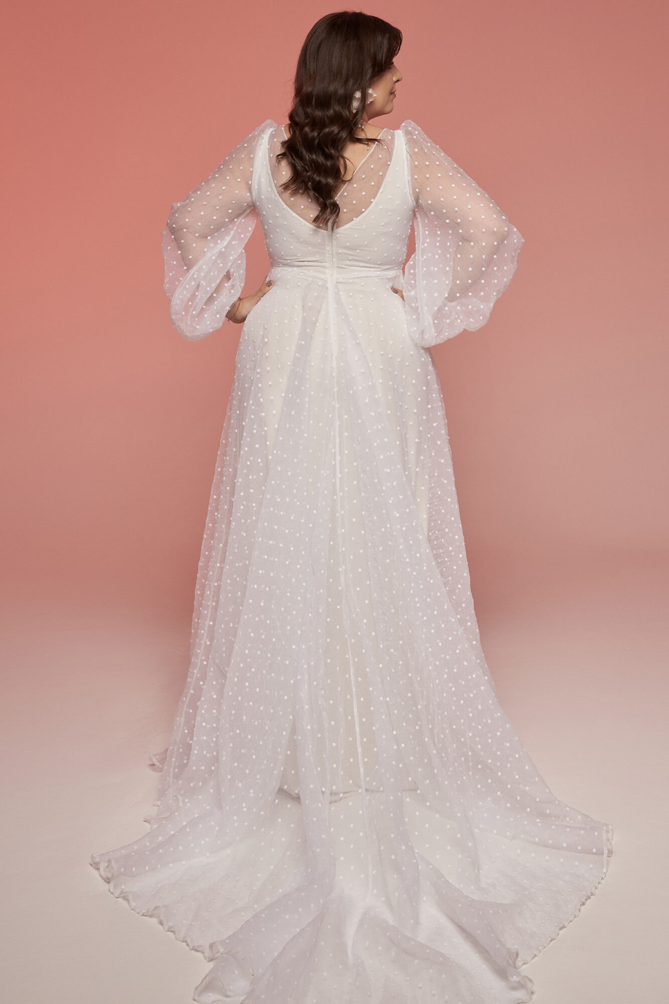 suknia ślubna z długimi, transparentnymi rękawami i koronkowym dołem, widoczna z tyłu na różowym tle