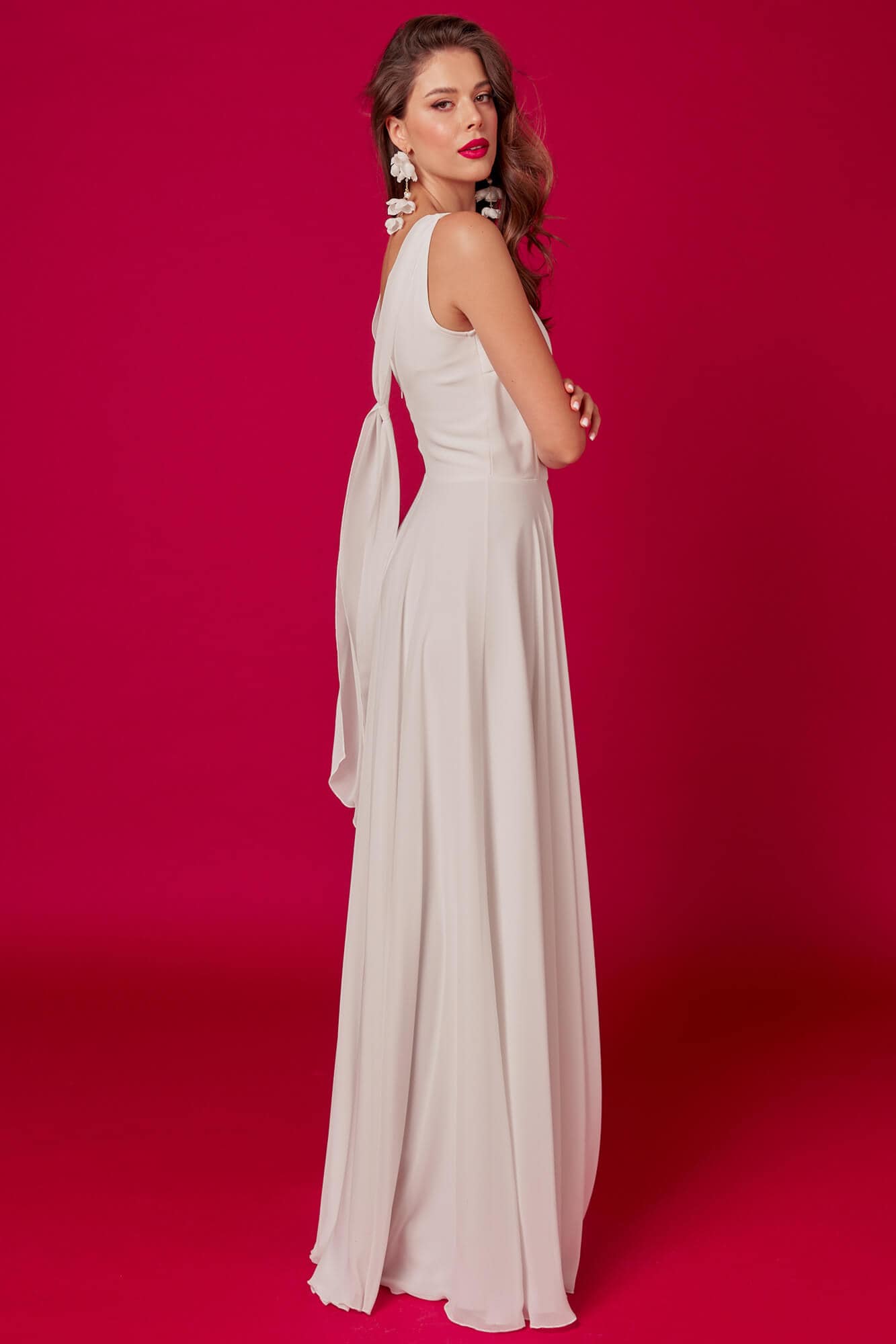 suknia ślubna w kolorze białym z długim tyłem i asymetrycznym dekoltem, modelka na tle różowej ściany