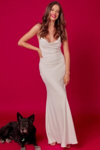 suknia w kolorze białym, obcisła, z dekoltem, modelka stoi obok czarnego psa na tle różowej ściany