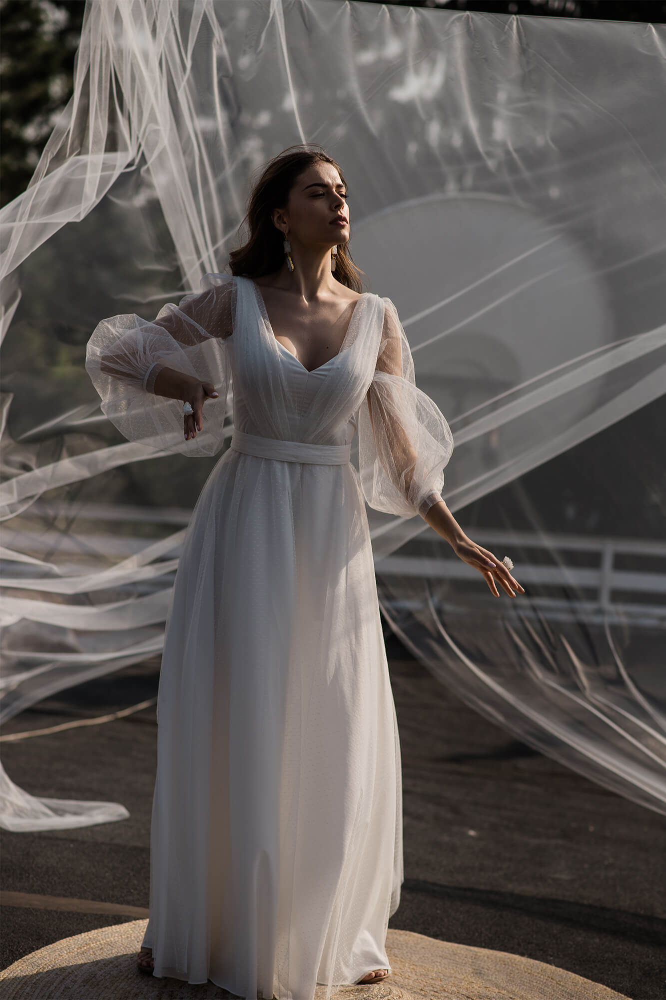suknia ślubna z długimi, przezroczystymi rękawami i dekoltem w serek, modelka na tle delikatnej tkaniny.