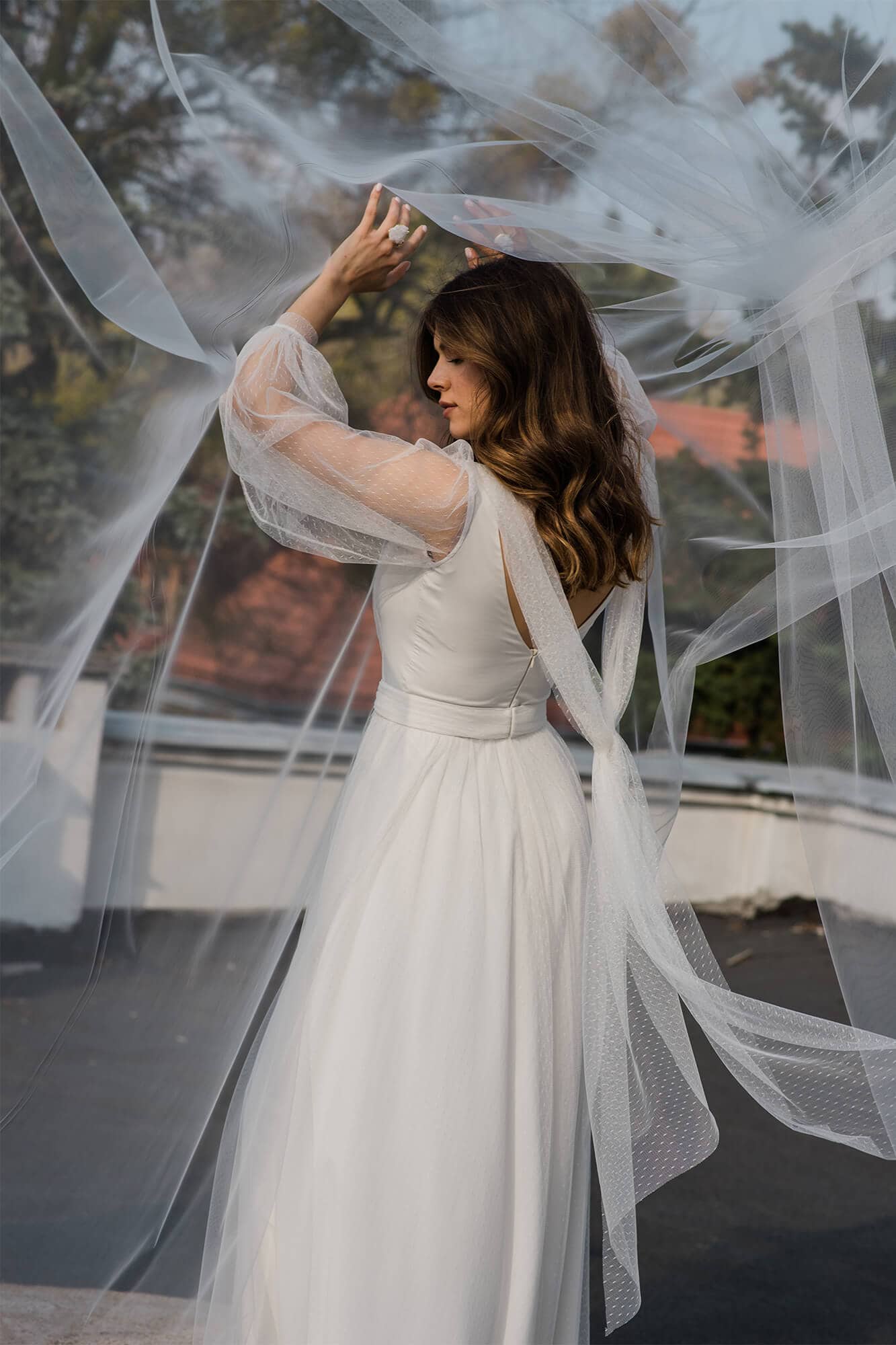 suknia ślubna z długimi rękawami i tiulowym welonem, modelka stoi na dachu budynku