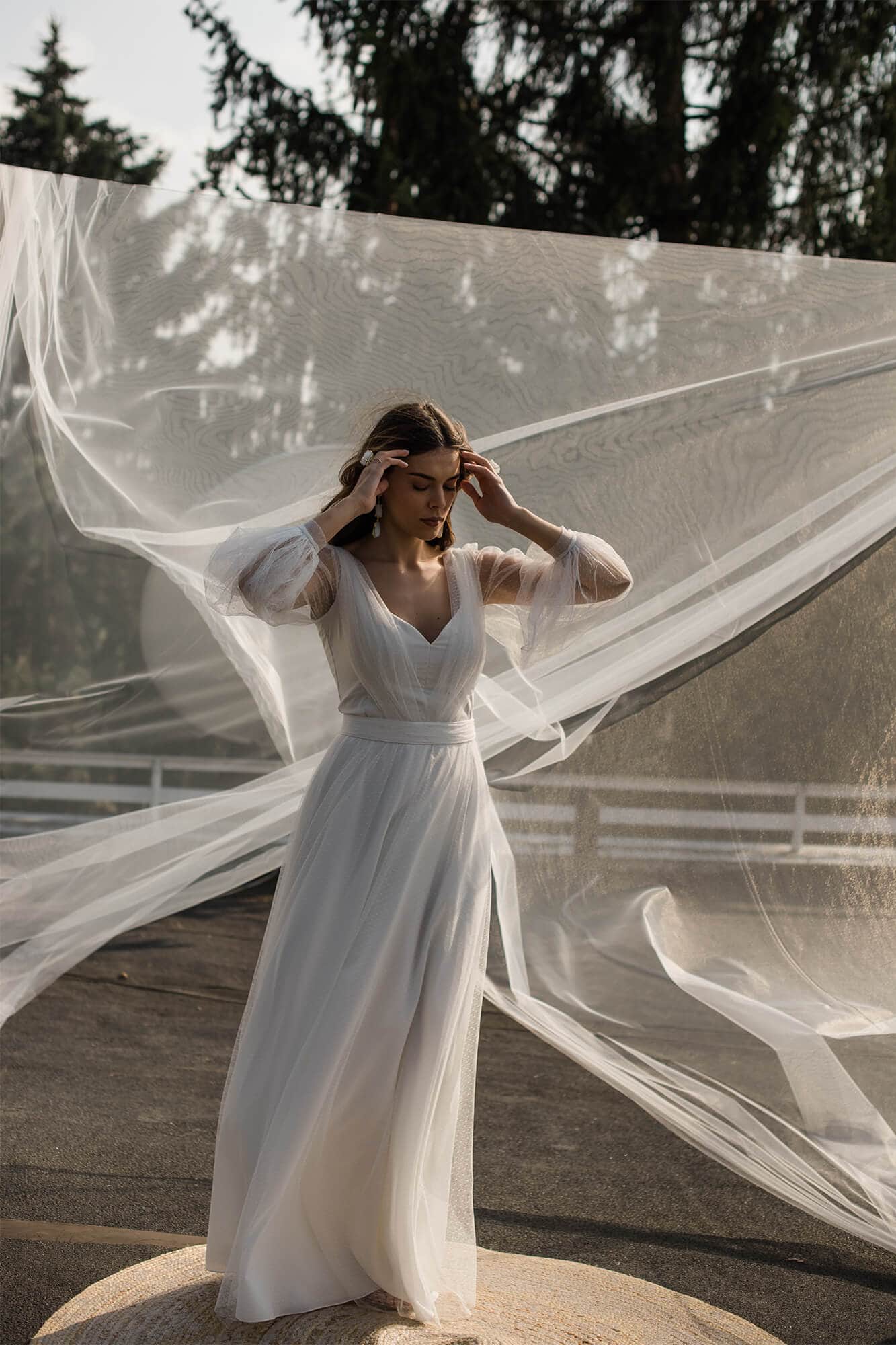 suknia ślubna z długimi rękawami, stojącą kobietą w tle i zwiewnym tiulem.