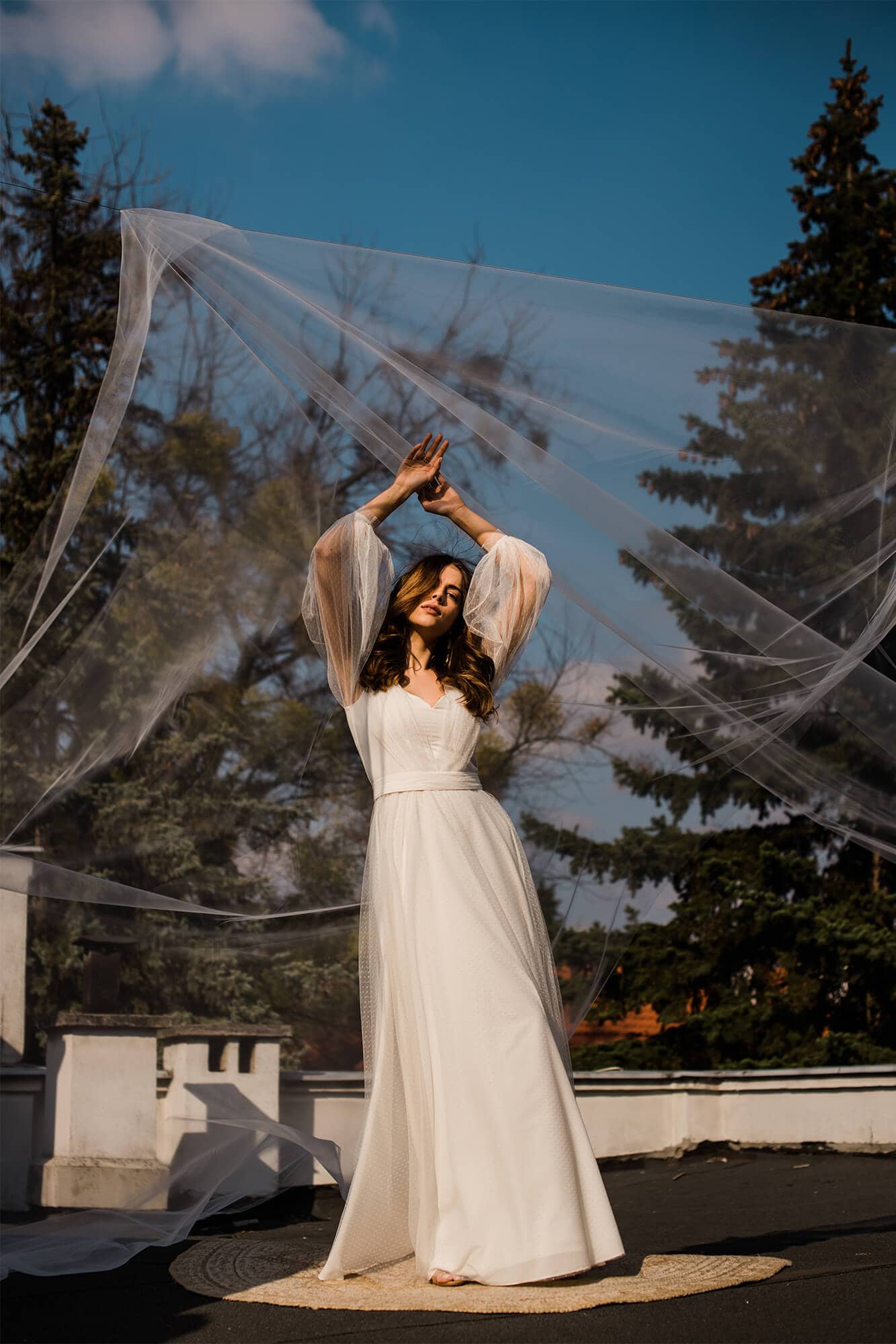 Suknia ślubna z długimi rękawami, kobieta w białym stroju, tło z drzewami i niebieskim niebem.
