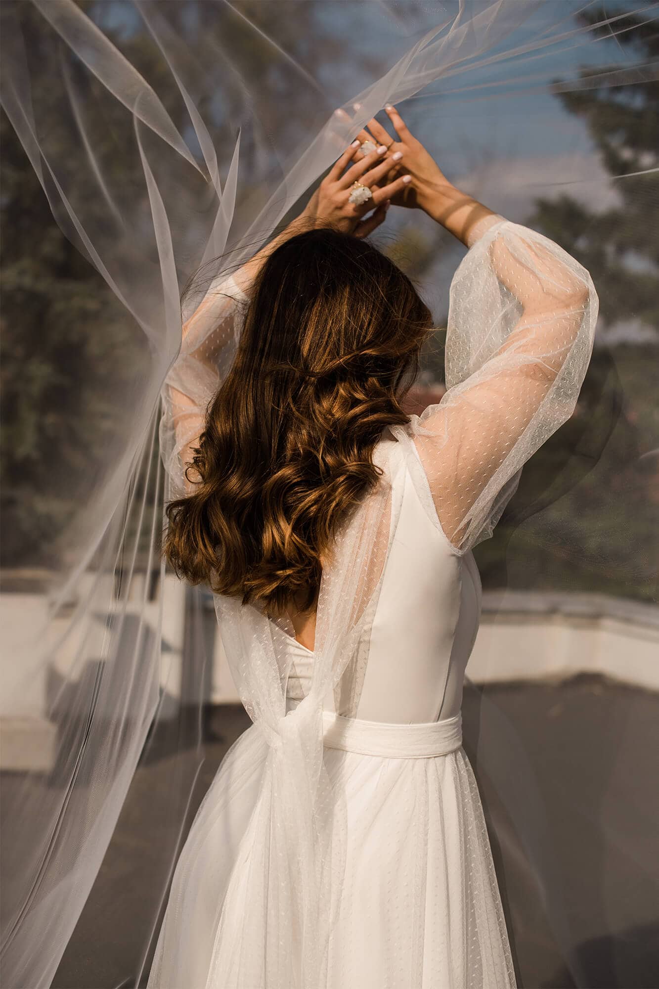 Suknia ślubna z długimi, przezroczystymi rękawami i wiązaniem w talii, z widocznym woalem.