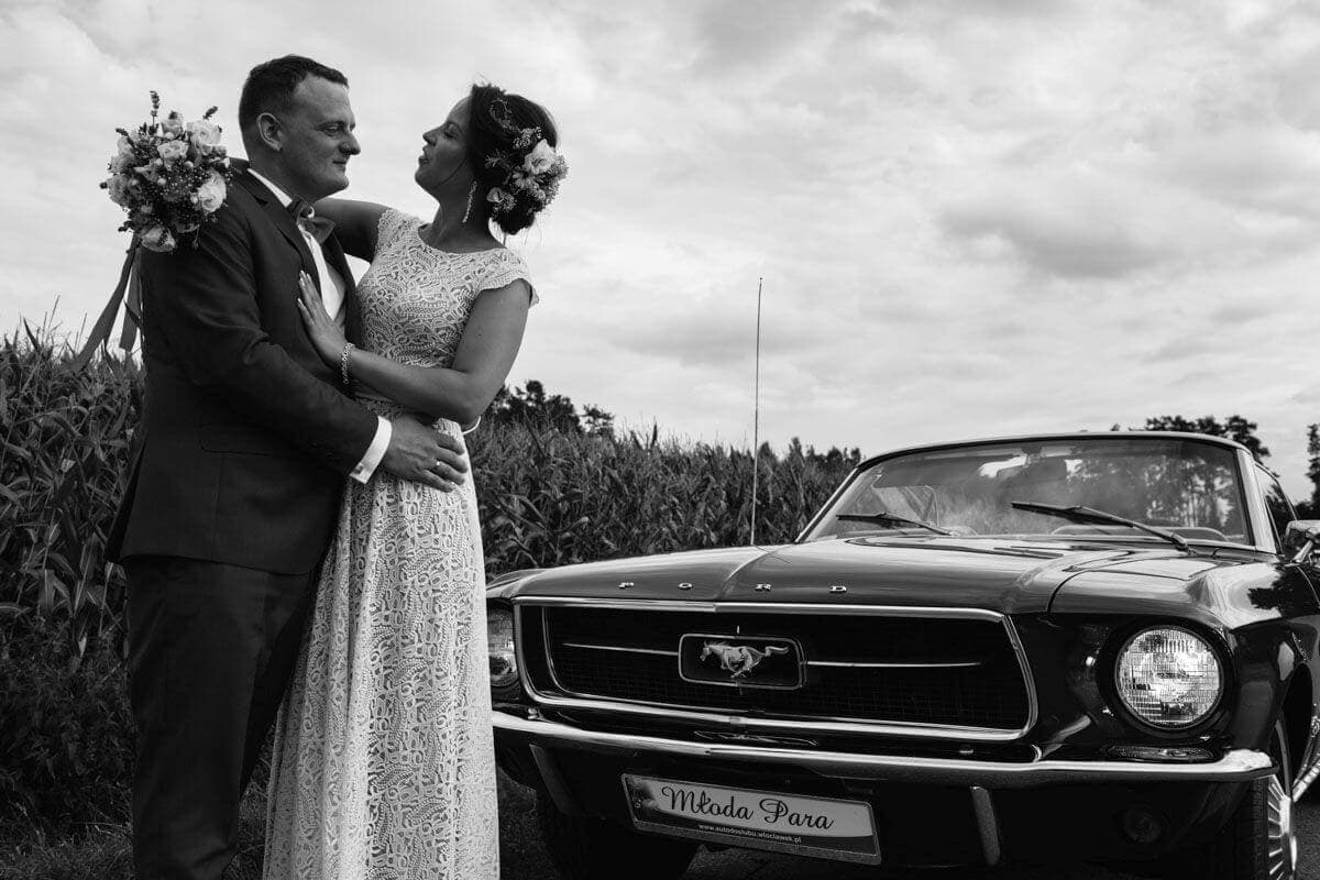 Para młoda w białej sukni ślubnej oraz garniturze stoi obok czarnego samochodu.