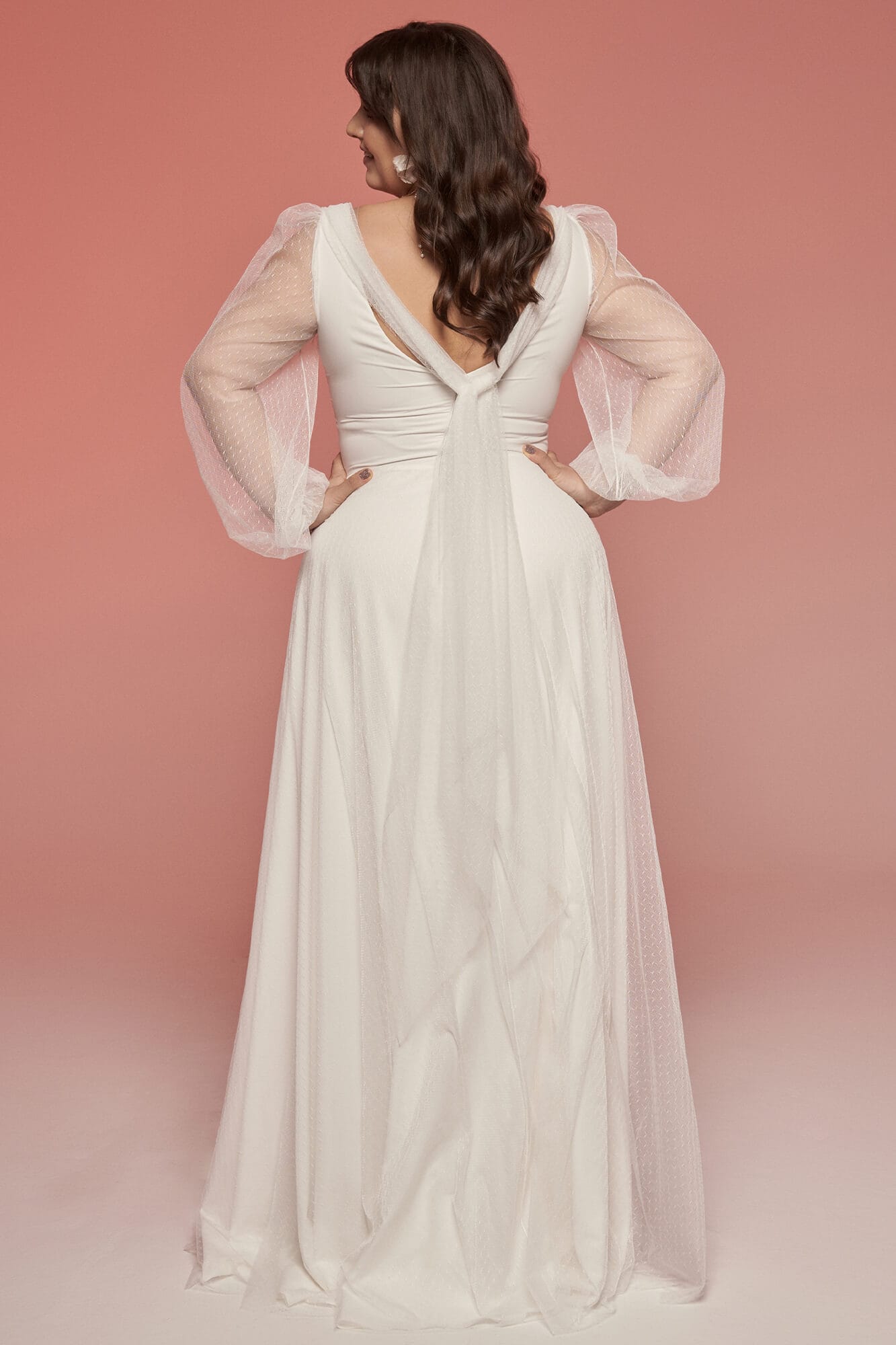 suknia ślubna z długimi, przezroczystymi rękawami i dekoltem w serek, widok z tyłu na różowym tle