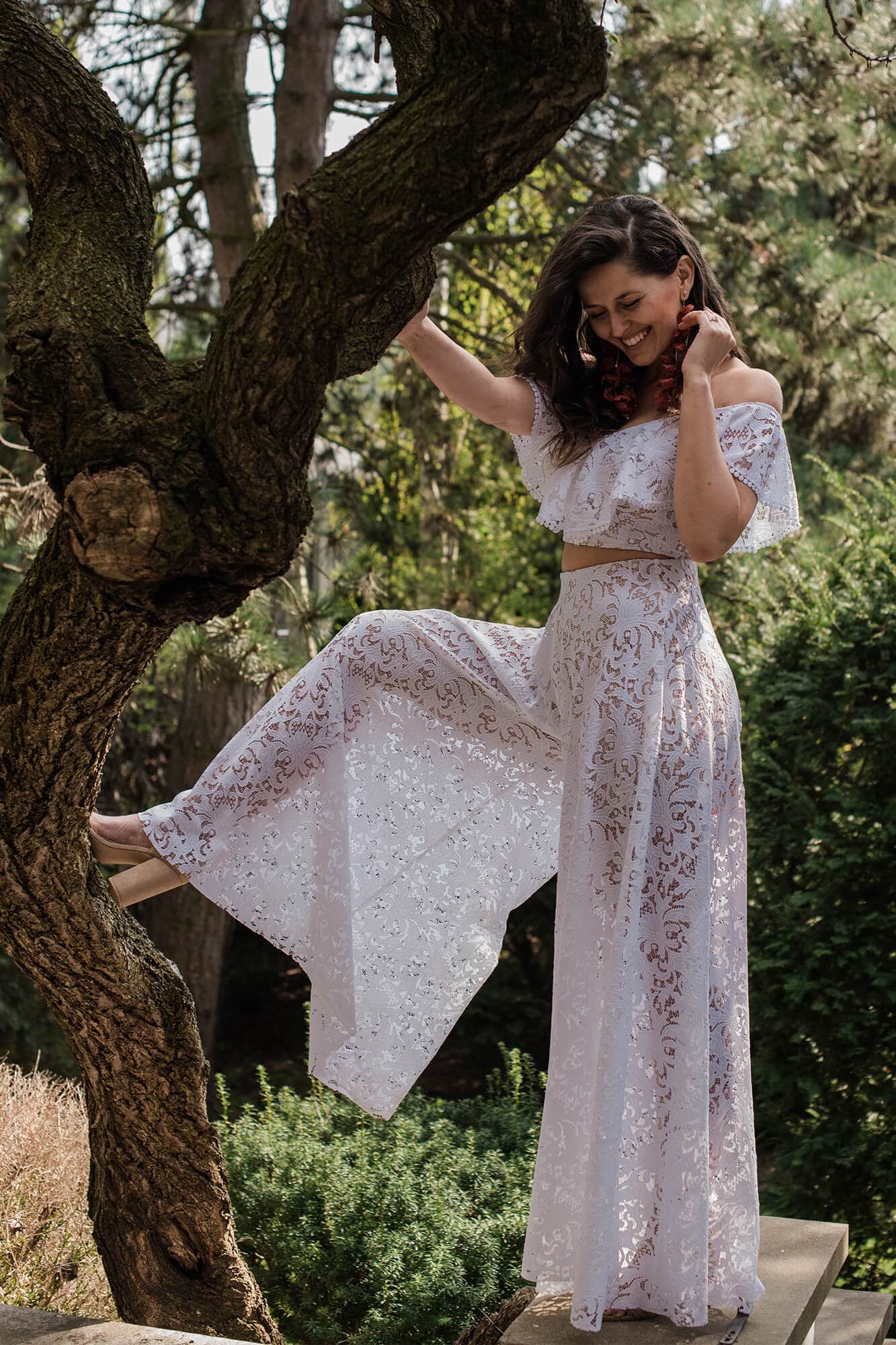 Suknia ślubna z koronkowymi detalami, z odkrytymi ramionami i szerokimi nogawkami, modelka stoi na drzewie.