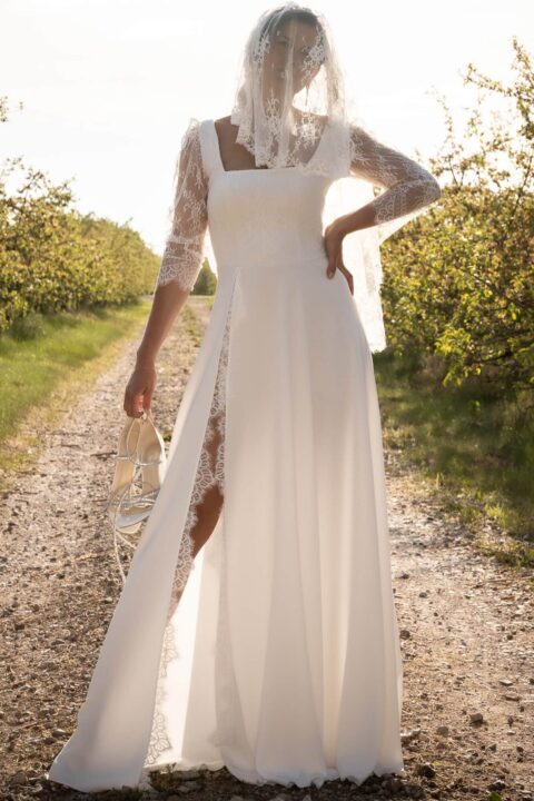 suknia ślubna z długimi koronkowymi rękawami i rozcięciem na boku, stojąca na ścieżce w otoczeniu zieleni