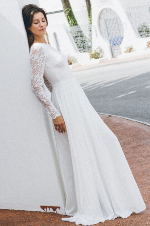 suknia ślubna z długimi koronkowymi rękawami i luźnym dołem, oparta o ścianę w plenerze
