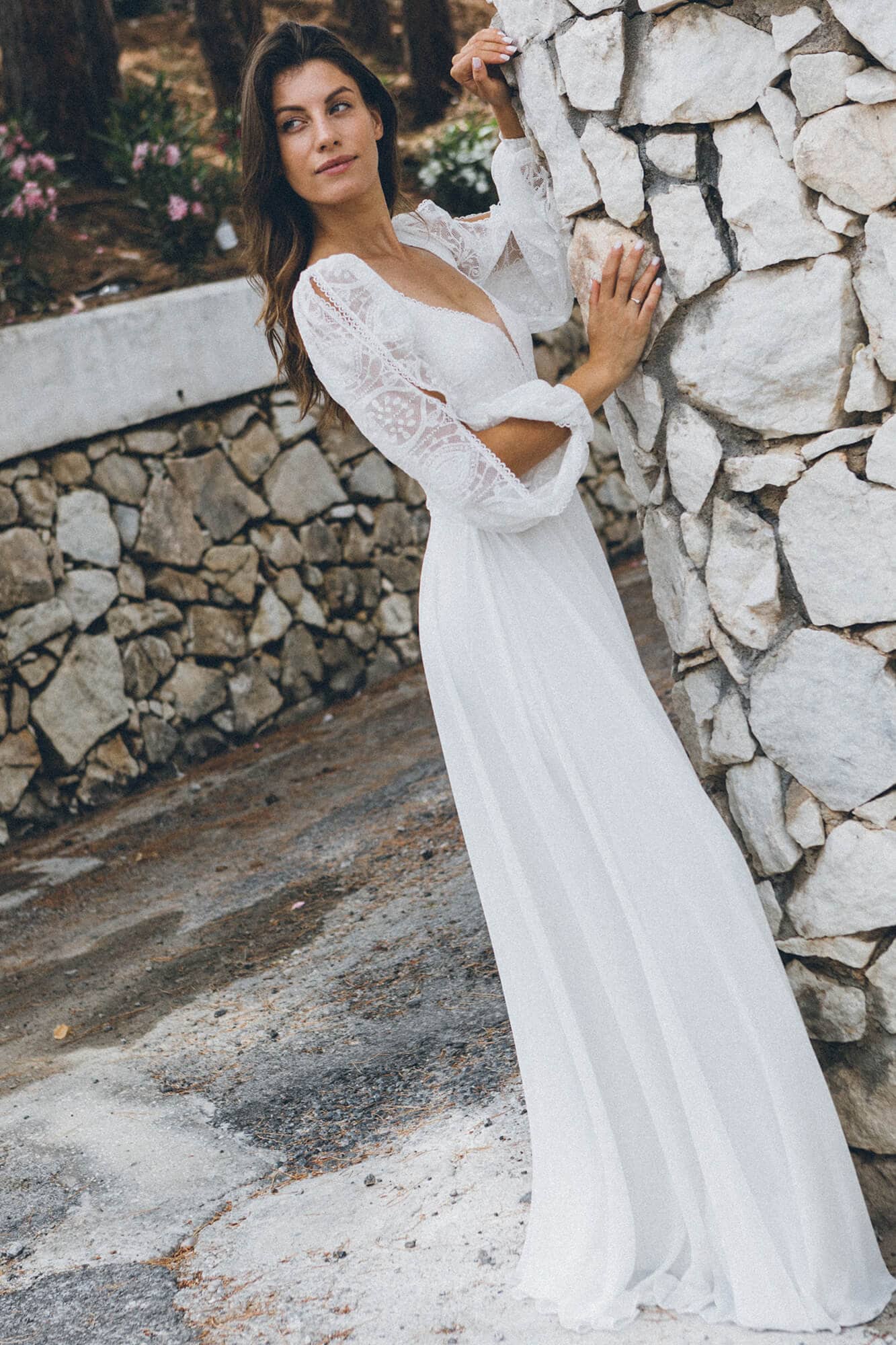 suknia ślubna z długimi koronkowymi rękawami, stojąca przy kamiennej ścianie w plenerze