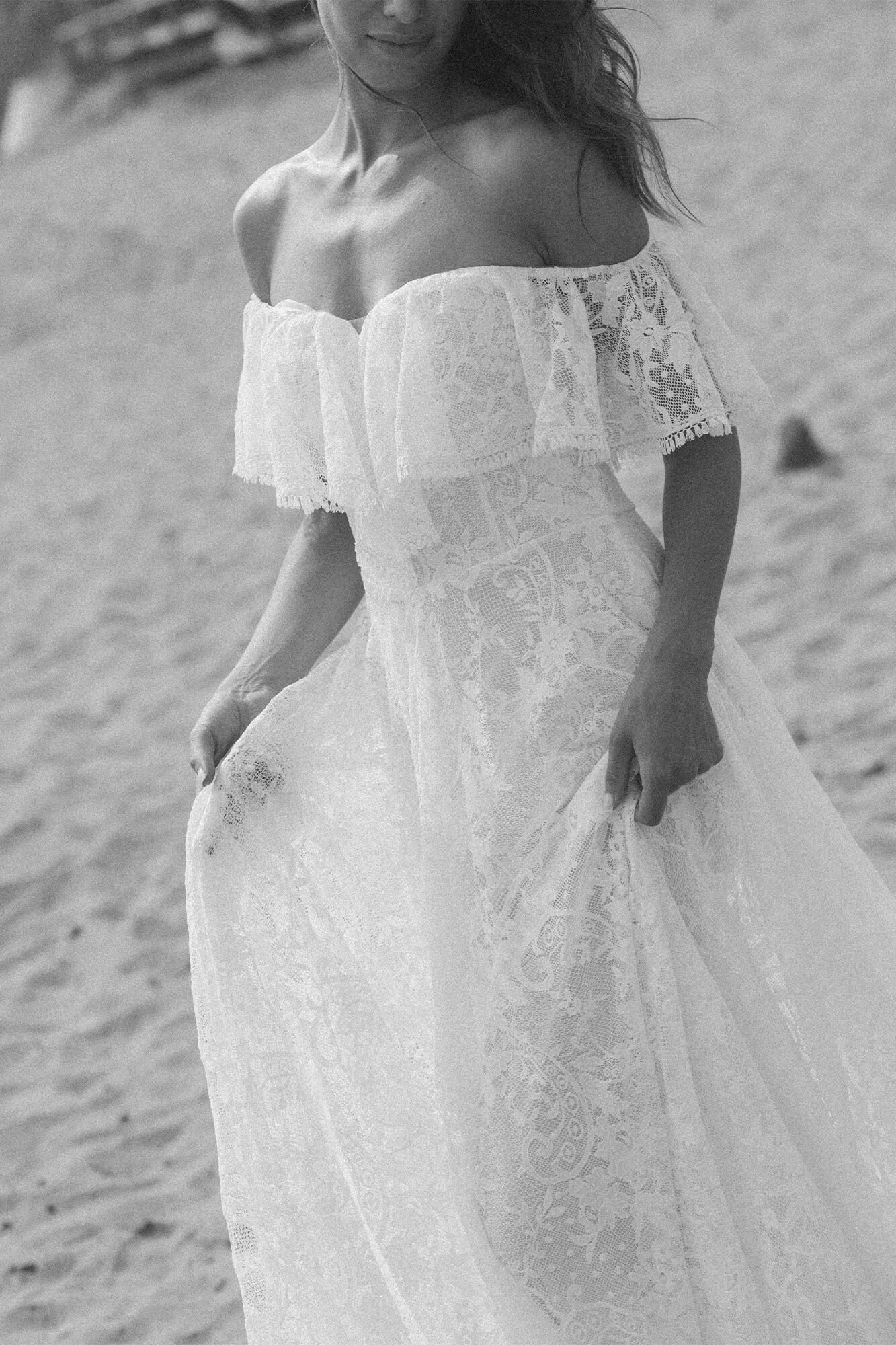 suknia ślubna z koronkowymi rękawami i falbaną, w kolorze białym, na tle piaszczystej plaży