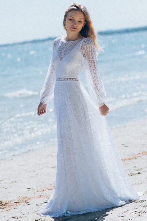 suknia ślubna z długimi rękawami, koronkowym wzorem i rozcięciem na dole, na tle plaży i morza