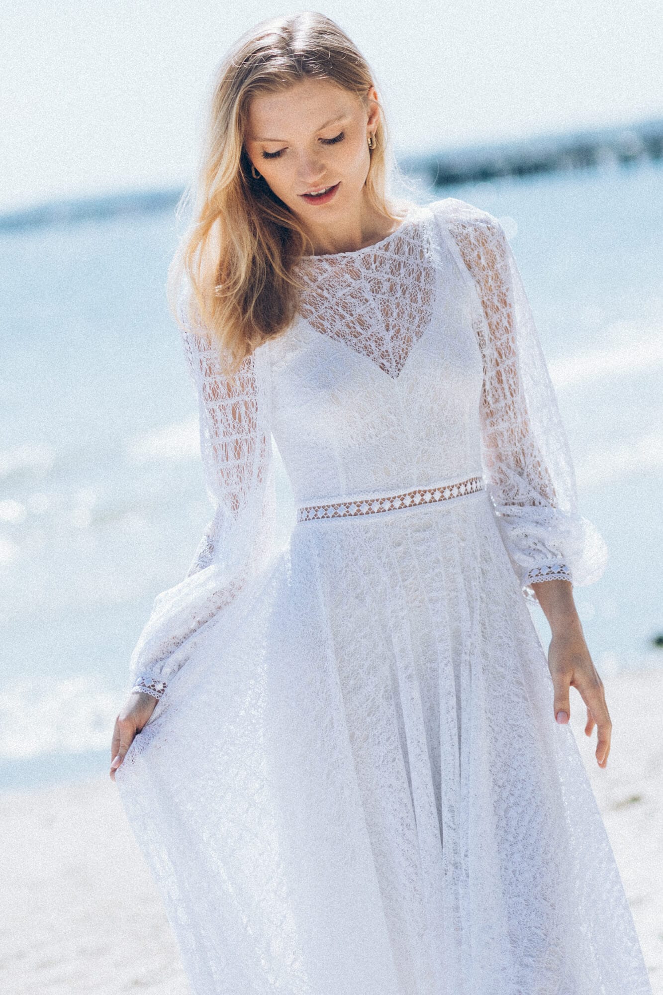 suknia ślubna z długimi rękawami, koronkowym wzorem i rozkloszowanym dołem, na tle plaży
