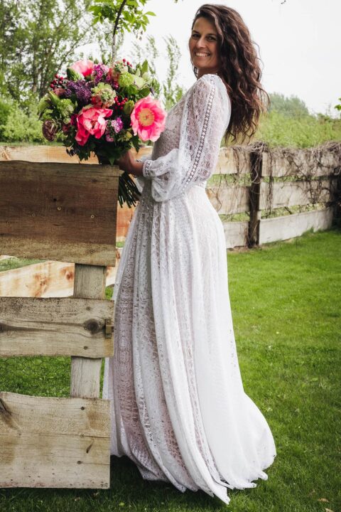 Suknia ślubna z długimi rękawami, widoczna na tle zielonej trawy i drewnianego ogrodzenia.