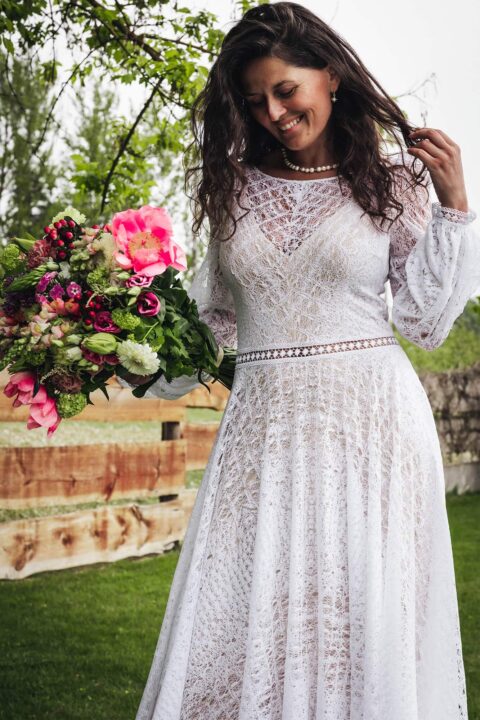 suknia ślubna z długimi rękawami, koronkowym wzorem i paskiem w talii, trzymana przez kobietę z bukietem kwiatów