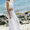 Suknia ślubna z koronkowym wykończeniem, z odkrytymi ramionami, na tle plaży i fal morskich.