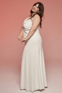 suknia ślubna w kolorze białym, z dekoltem w serek i długim krojem, modelka stoi na tle różowej ściany