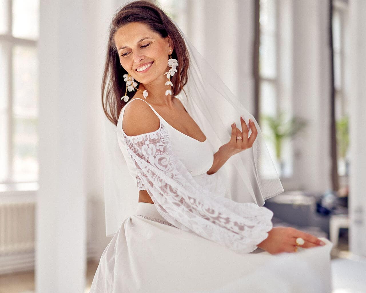 Kobieta w białej sukni ślubnej z koronkowymi rękawami i welonem, uśmiechająca się w jasnym wnętrzu.