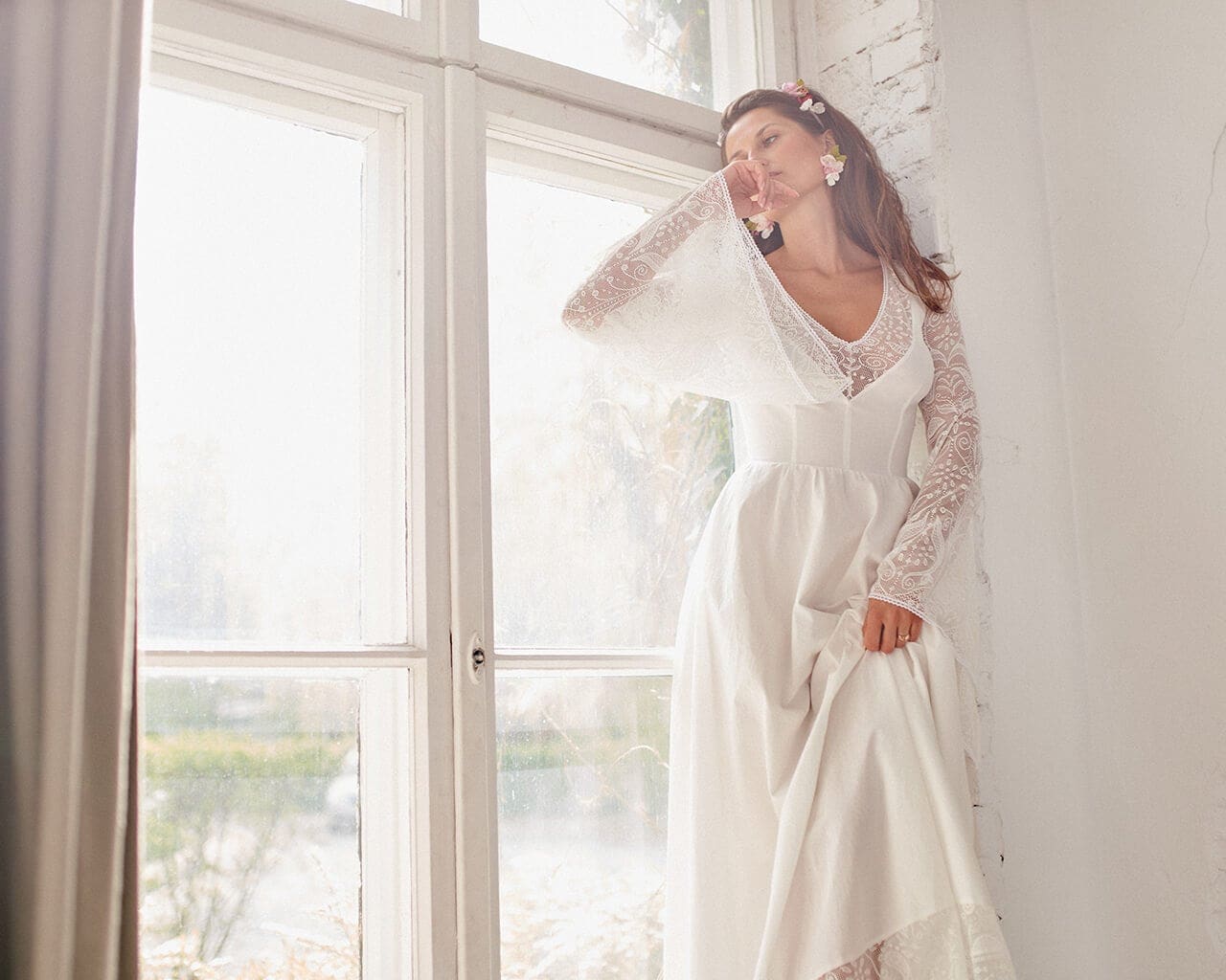 suknia ślubna z długimi koronkowymi rękawami i dekoltem w serek, modelka stoi przy oknie.
