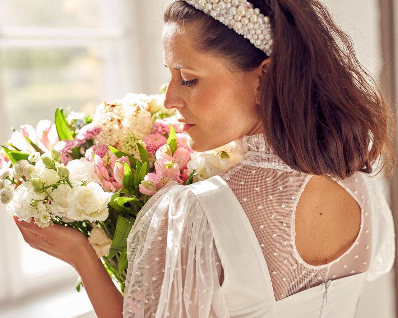 Kobieta trzyma bukiet kwiatów, ubrana w suknię ślubną z przezroczystymi rękawami.