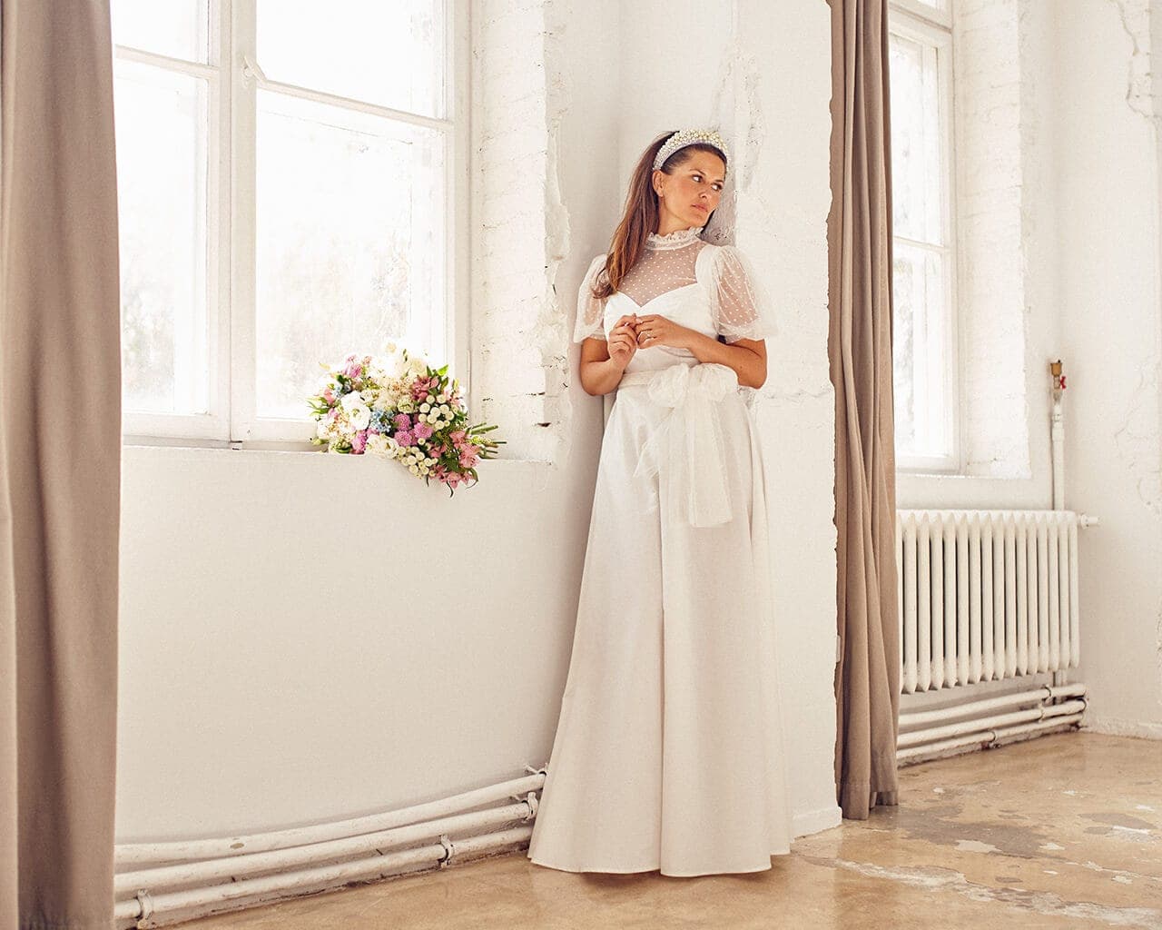 Suknia ślubna z długą spódnicą i koronkowym topem, modelka stoi obok okna z bukietem kwiatów.
