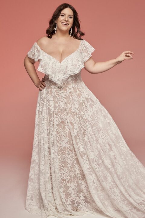 suknia ślubna z koronkowym wzorem i falbaniastymi rękawami, na modelce stojącej na tle w kolorze różowym