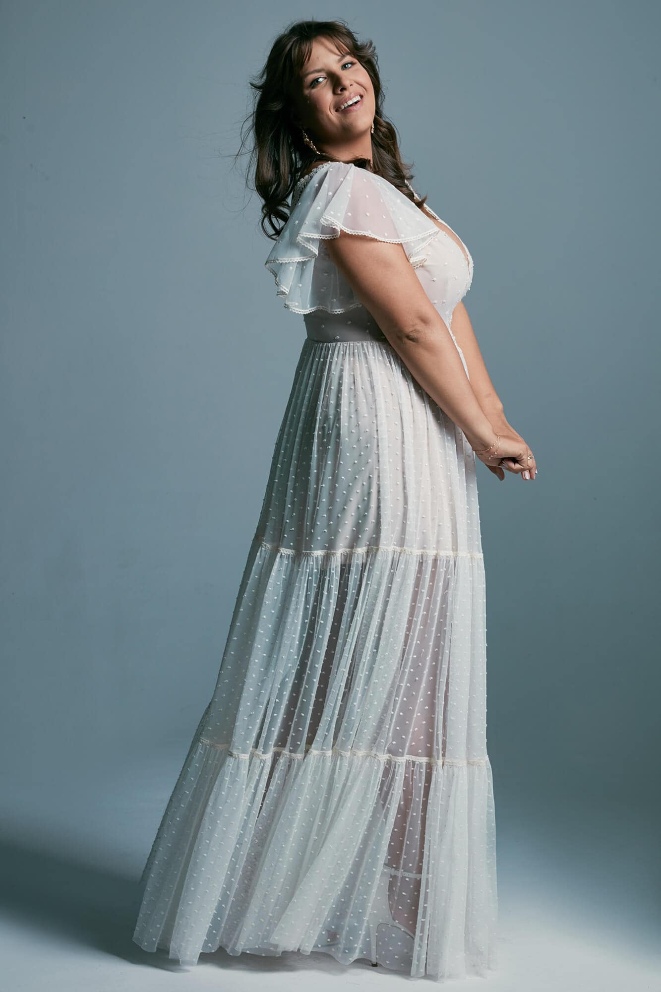 suknia ślubna z przezroczystymi wstawkami i falbaniastymi rękawami, modelka w profilu na tle szarym