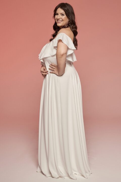 suknia ślubna w kolorze białym z odkrytymi ramionami i falbanami, modelka w pozycji bocznej na różowym tle