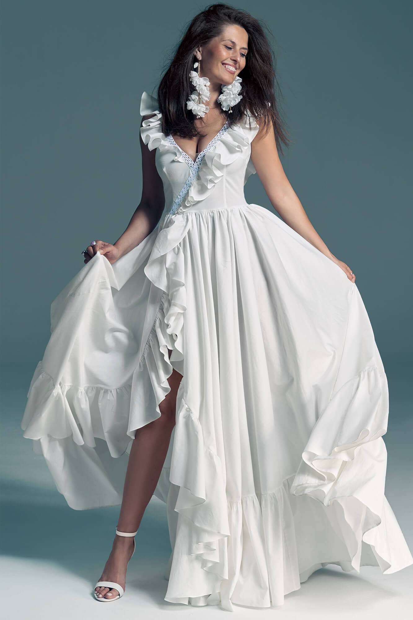 suknia ślubna z falbanami, dekoltem w serek i rozcięciem na boku, prezentowana przez kobietę w białych sandałach