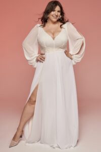 suknia ślubna z długimi rękawami, dekoltem w serek i rozcięciem z boku, noszona przez kobietę na różowym tle