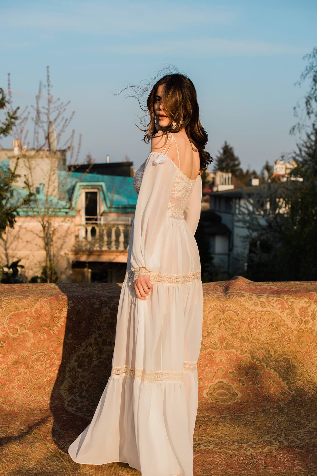 suknia w kolorze białym z długimi rękawami, stojąca na tle budynków i roślinności