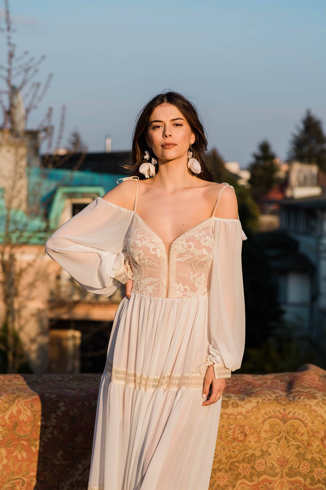 Suknia z długimi rękawami, dekoltem w serek i koronkowymi detalami, modelka na tle budynków.