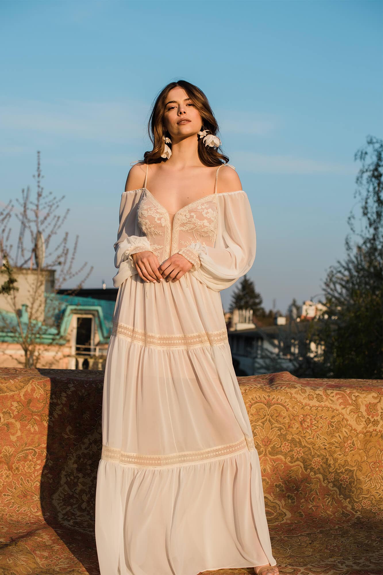 suknia ślubna w kolorze kremowym z koronkowymi detalami i długimi rękawami, modelka stoi na tle nieba