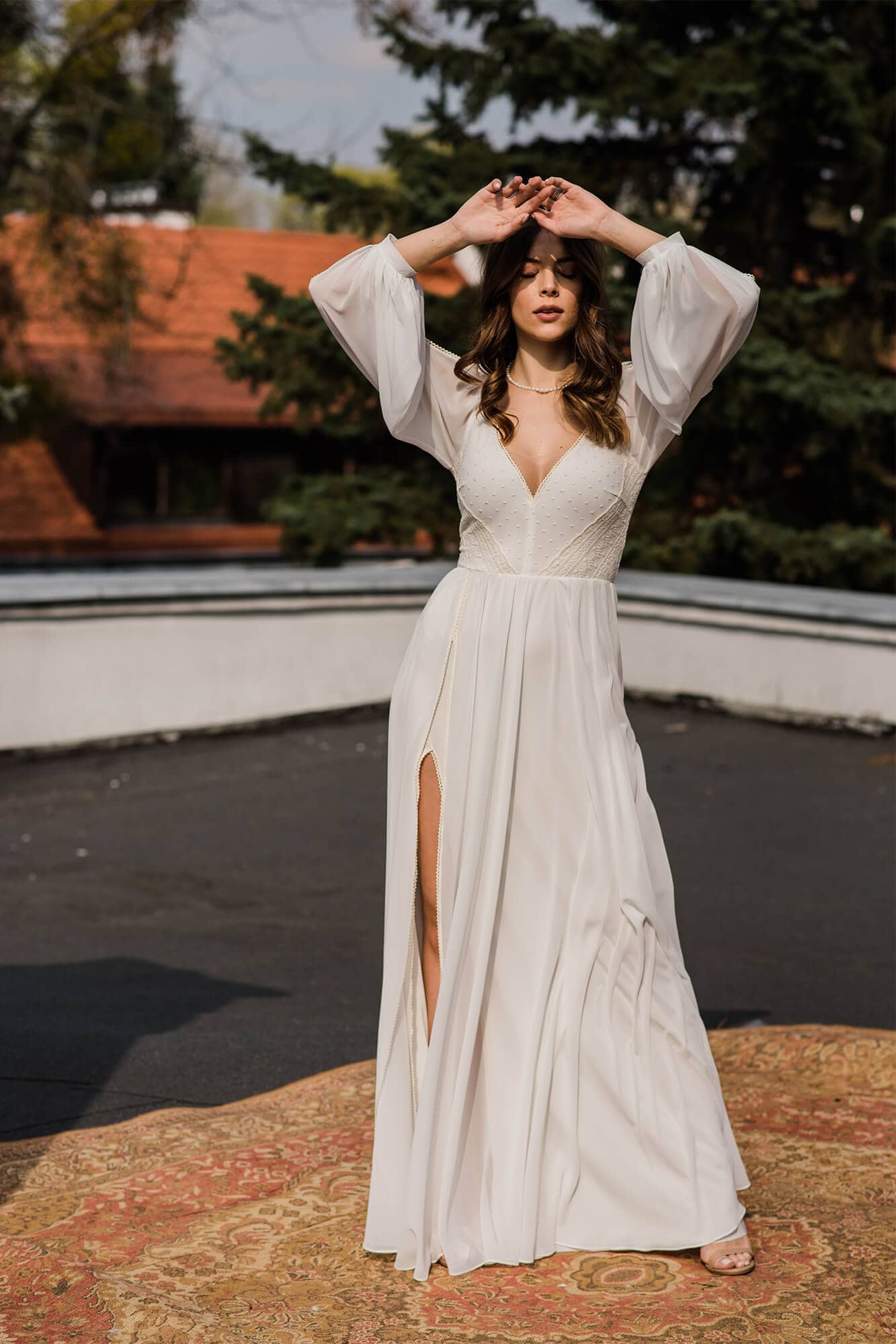 suknia ślubna z długimi rękawami i rozcięciem po boku, modelka na tle budynku.