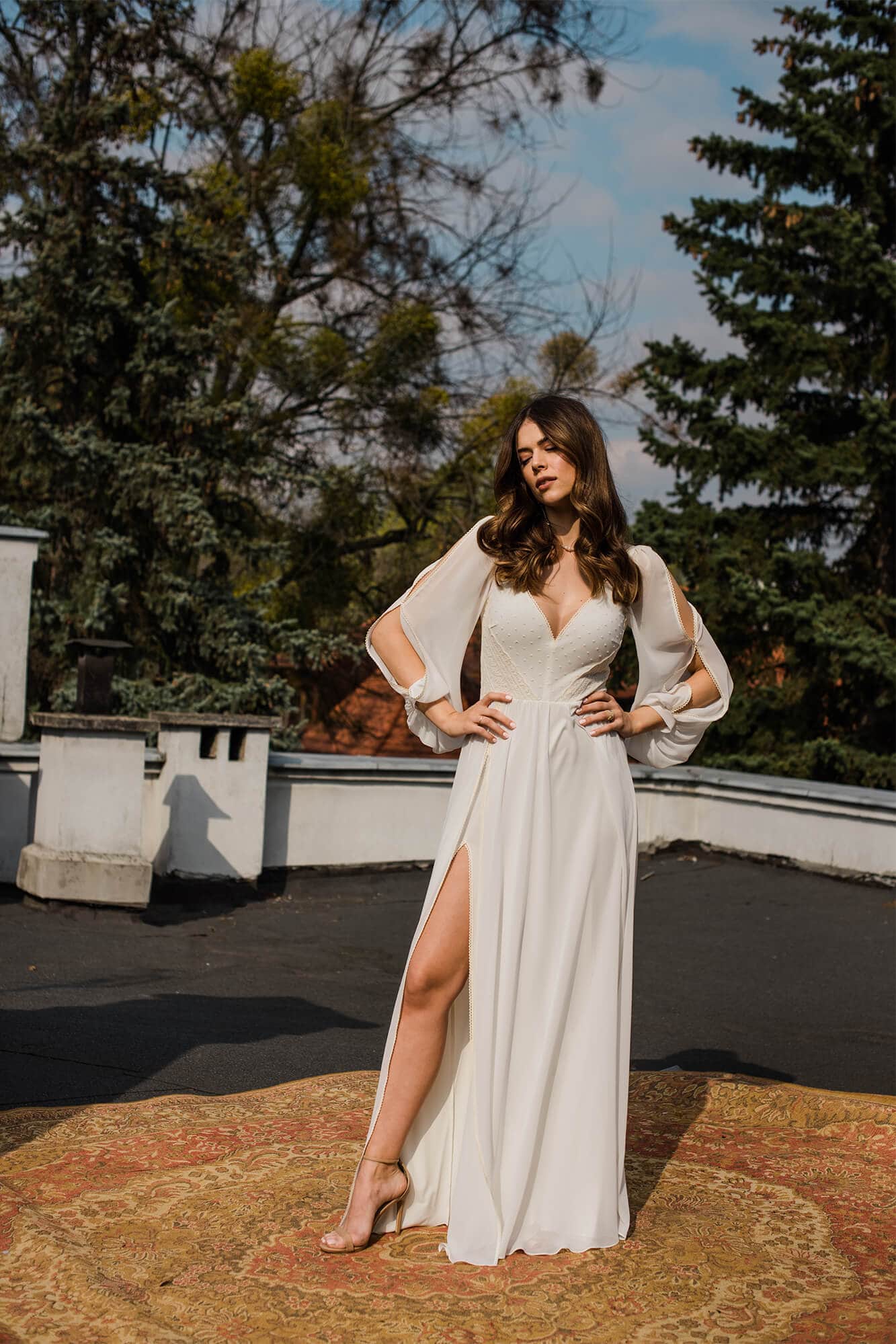 suknia ślubna z głębokim dekoltem, długimi rękawami i rozcięciem na nodze, na tle zieleni i nieba