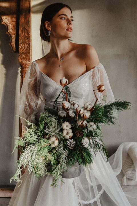 suknia ślubna z dekoltem w serek i koronkowymi rękawami, trzymana przez osobę z bukietem kwiatów