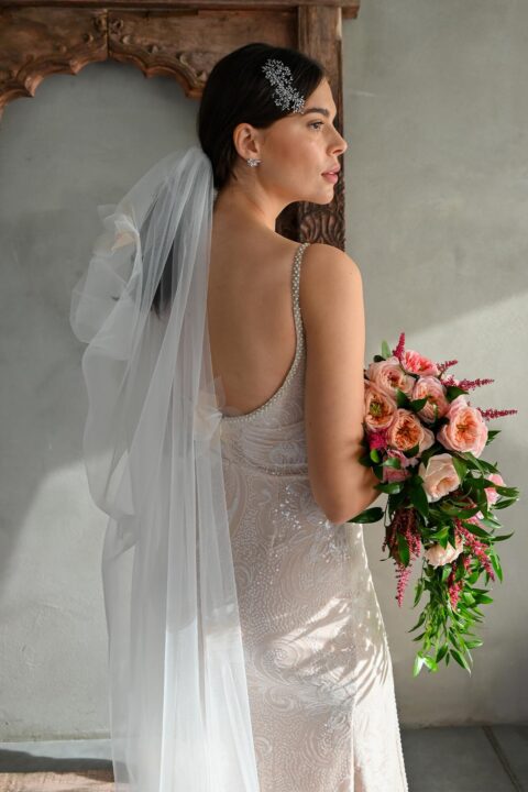suknia ślubna z koronkowymi detalami, długim welonem i bukietem róż w dłoni modelki