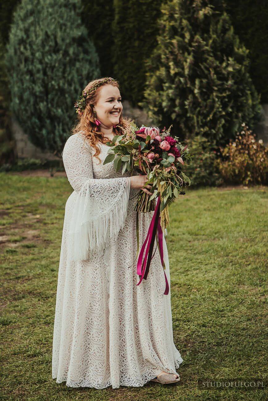 Kobieta w długiej sukni ślubnej trzyma bukiet kwiatów na tle zielonej trawy.