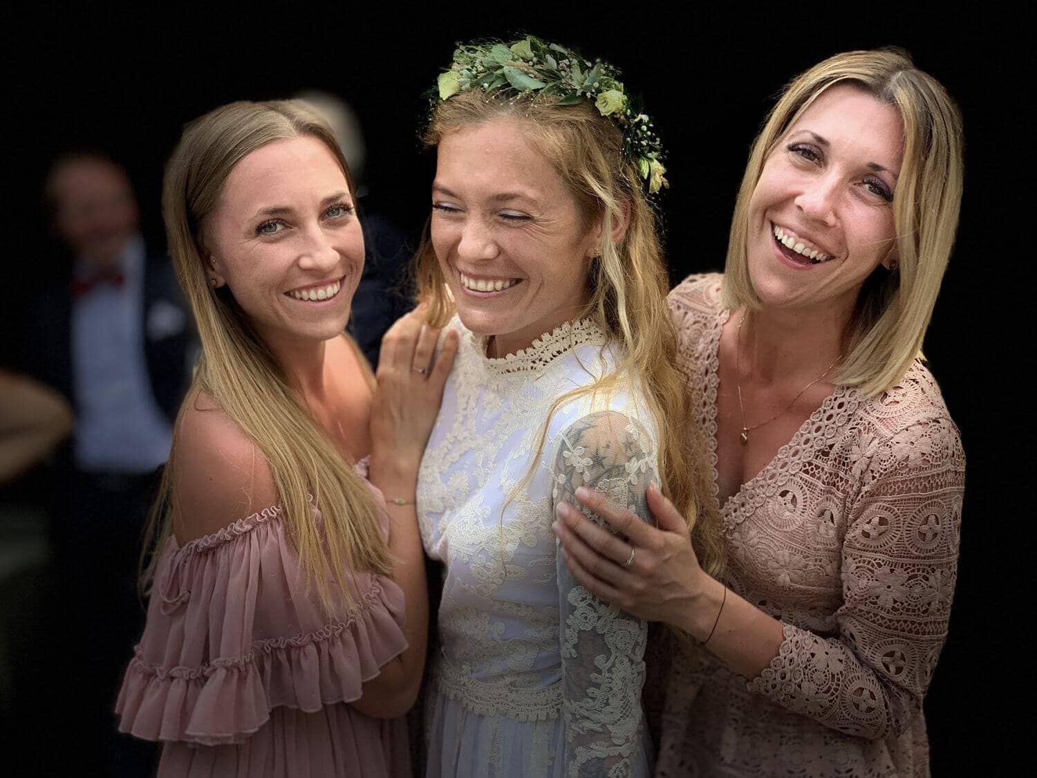Trzy kobiety uśmiechają się, jedna z nich ma na sobie białą suknię i wianek na głowie.