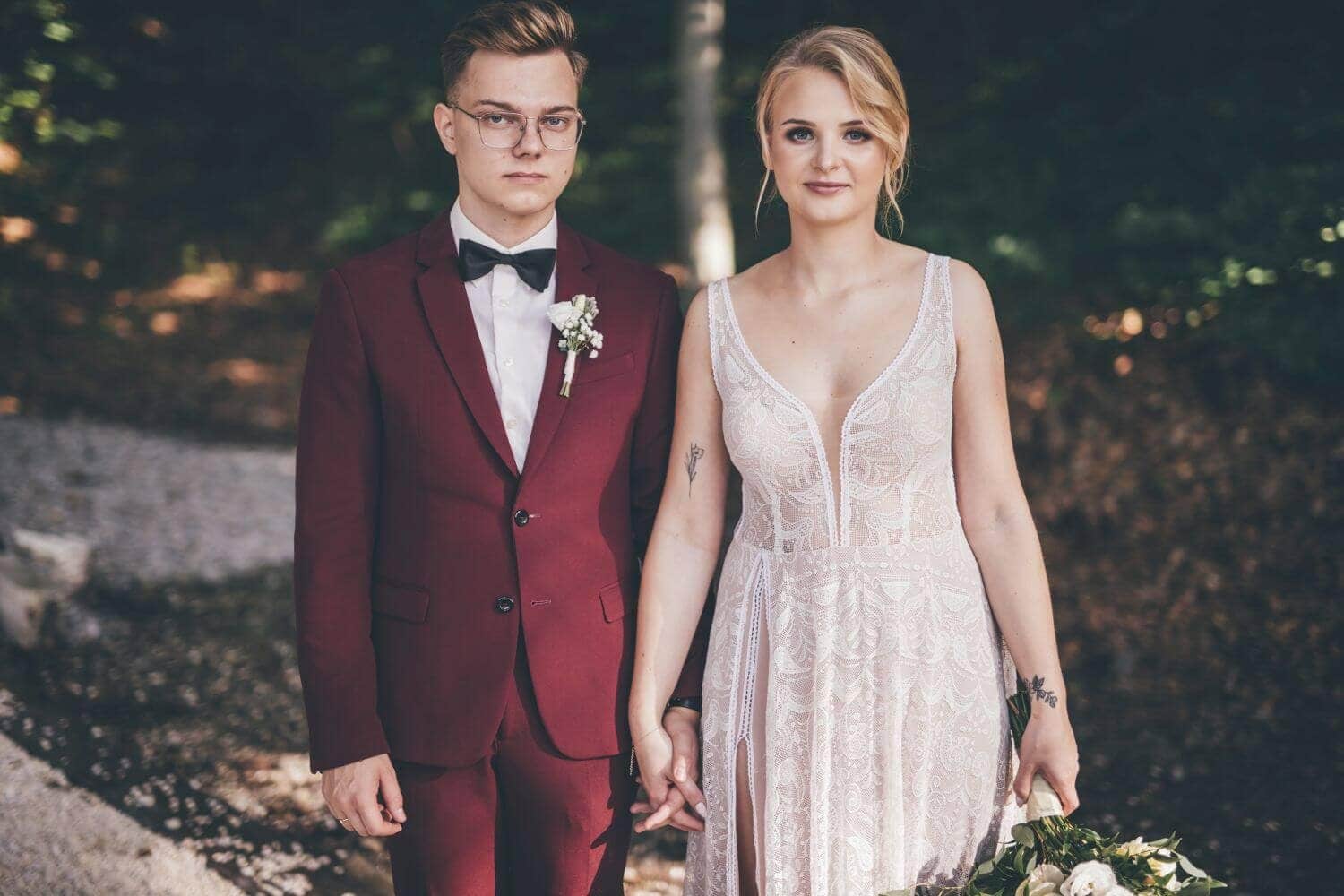 Osoba w sukni ślubnej z koronkowym wzorem trzymającą bukiet, obok mężczyzna w burgundowym garniturze.