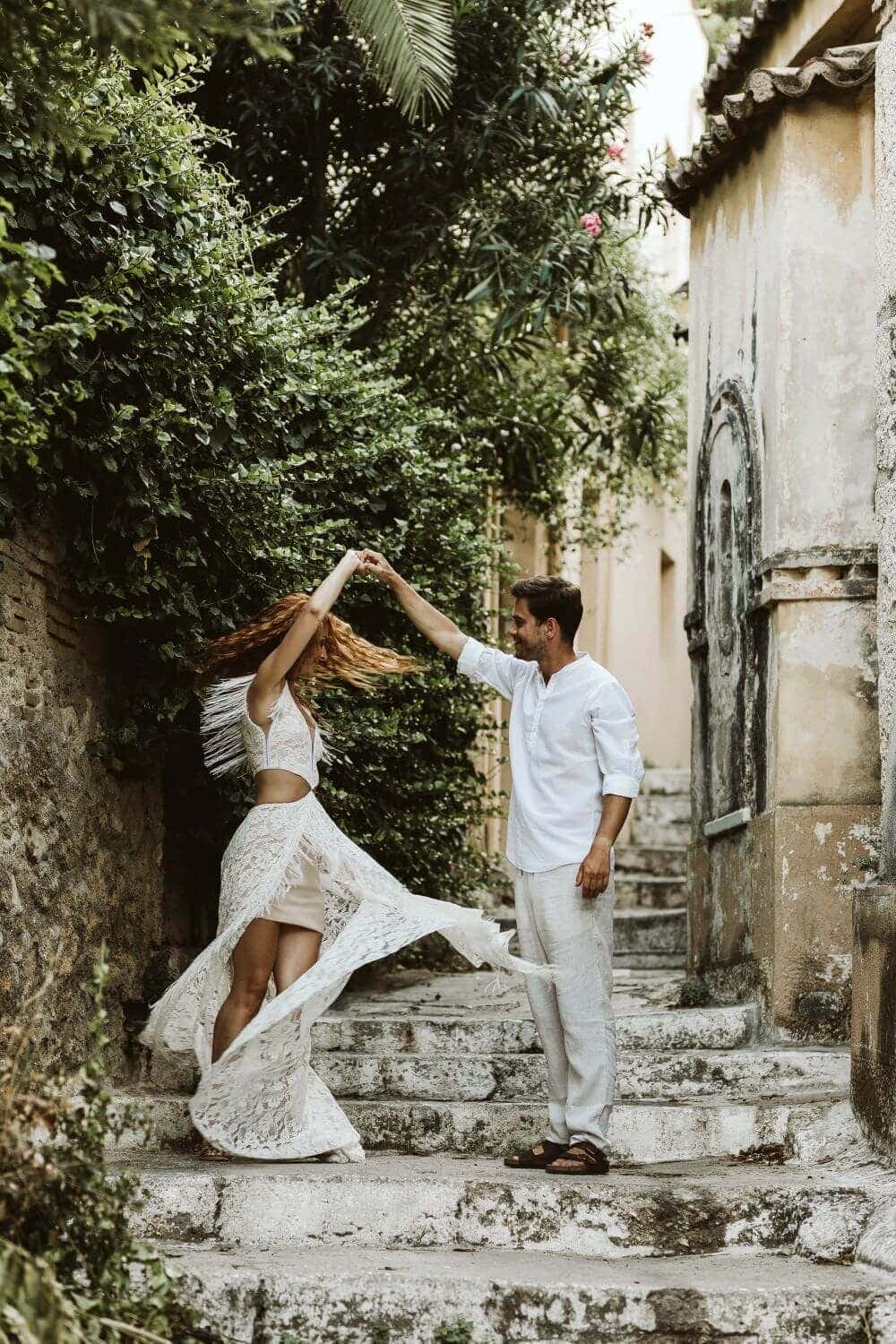 Osoba w białej sukni tańczy z mężczyzną w białej koszuli na schodach w otoczeniu zieleni.