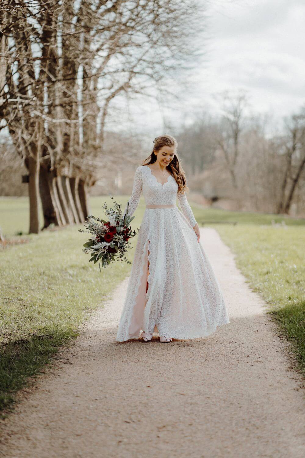 Kobieta w długiej sukni ślubnej trzymająca bukiet, spacerująca po ścieżce w parku.