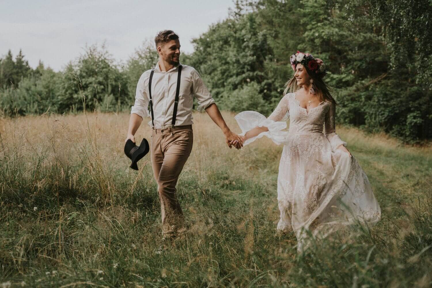 Osoba trzymająca w ręku kapelusz spaceruje z kobietą w sukni ślubnej po zielonej łące.