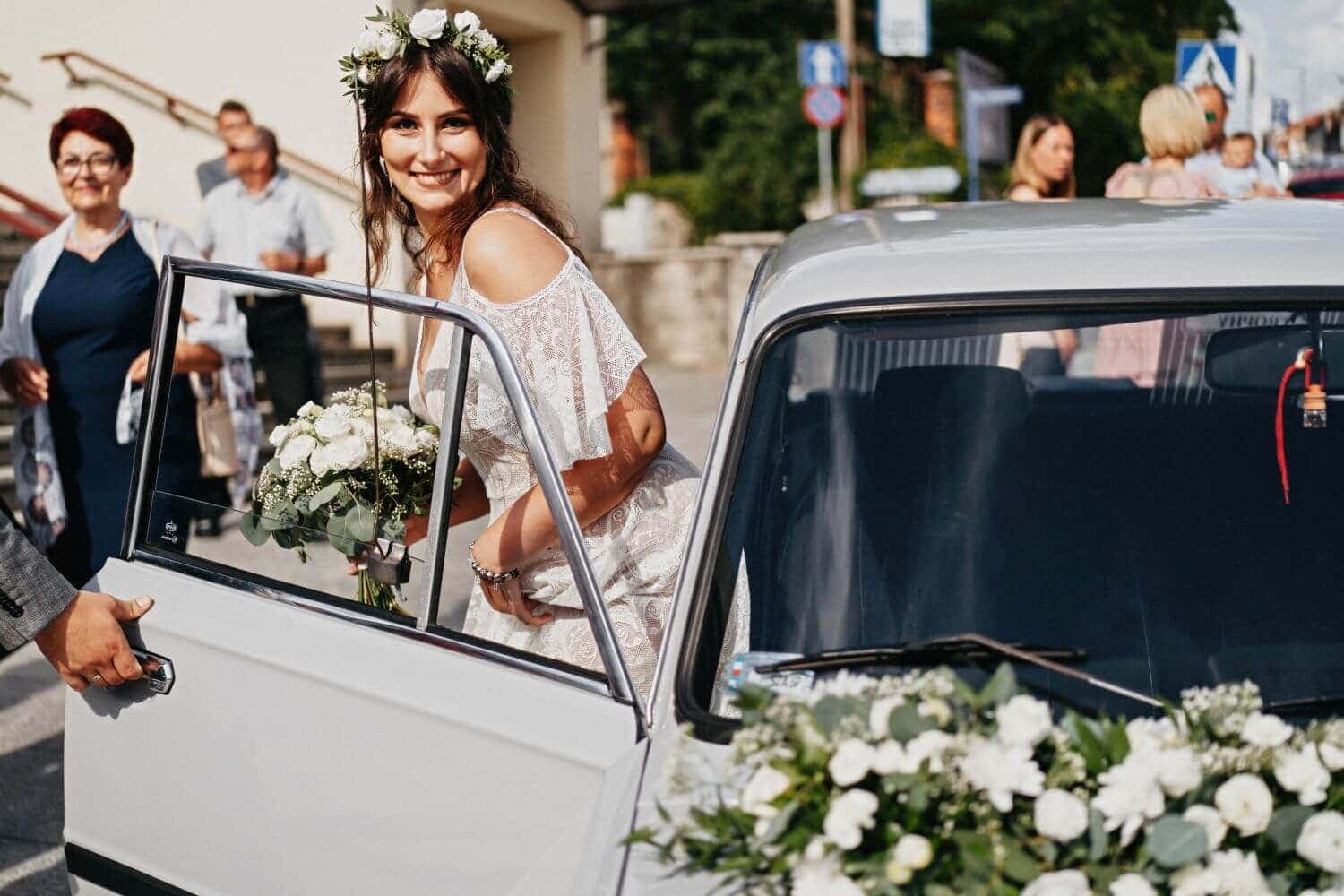 Kobieta w białej sukni z kwiatami we włosach wchodzi do samochodu ozdobionego kwiatami.
