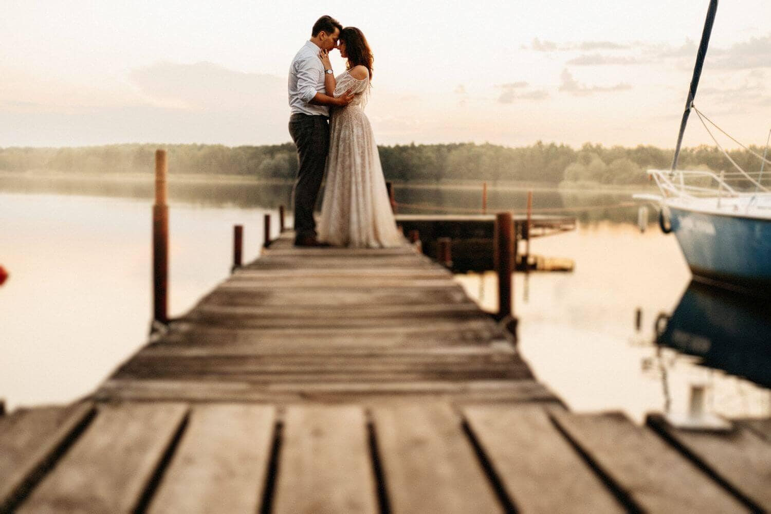 Para stoi na drewnianym pomoście nad wodą, kobieta w długiej sukni ślubnej.