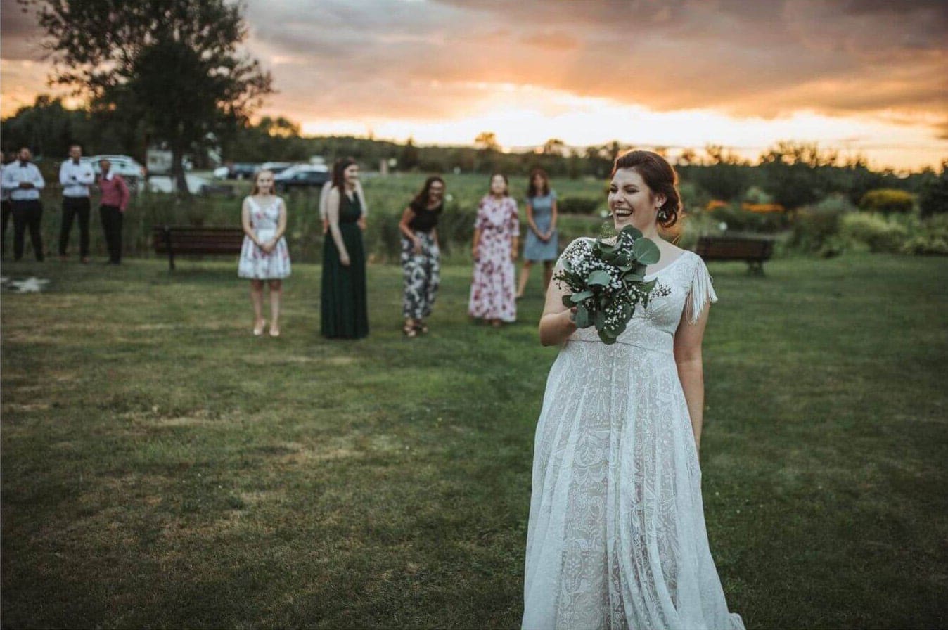 Suknia ślubna z koronkowym wykończeniem, kobieta trzyma bukiet, w tle goście na polanie.
