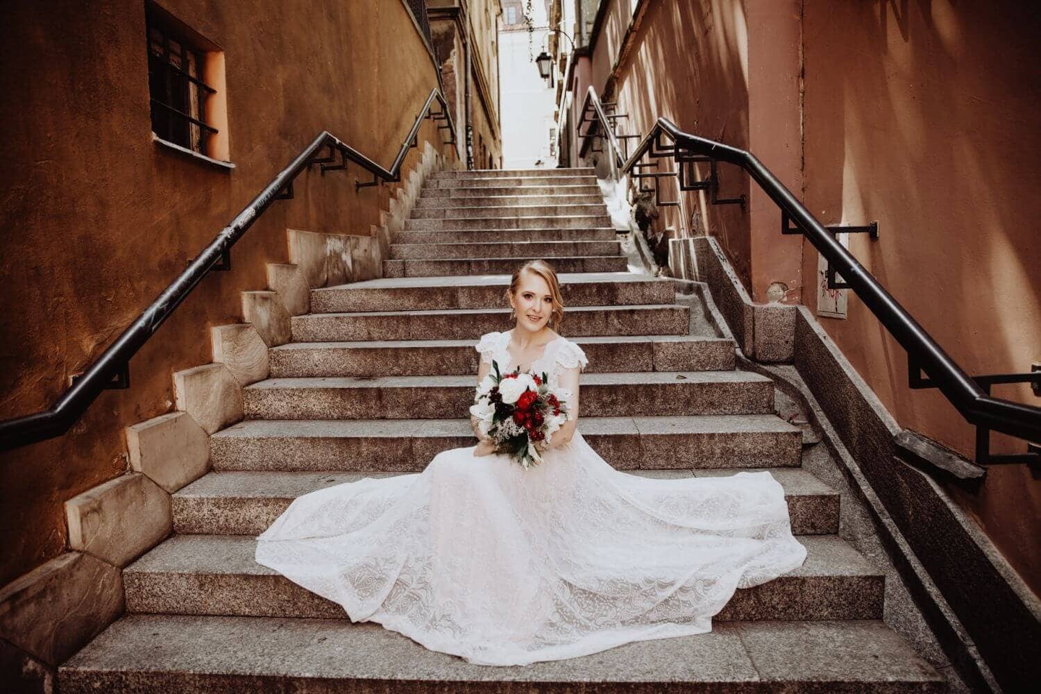 Kobieta w sukni ślubnej siedzi na schodach, trzymając bukiet kwiatów.