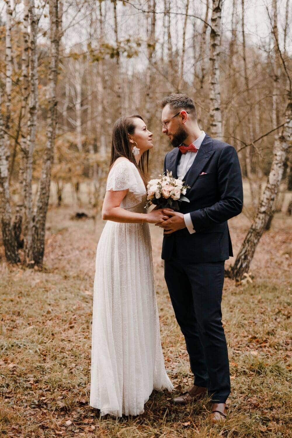 Para w eleganckich strojach stoi w lesie, kobieta trzyma bukiet kwiatów.