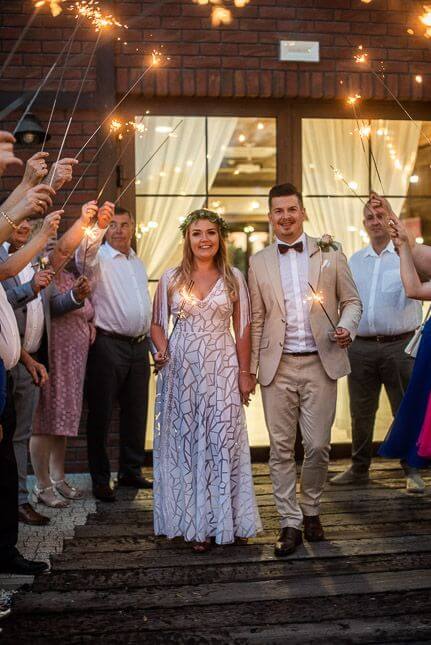 Para młoda trzymająca się za ręce, otoczona przez gości trzymających raczki na tle jasnych drzwi.